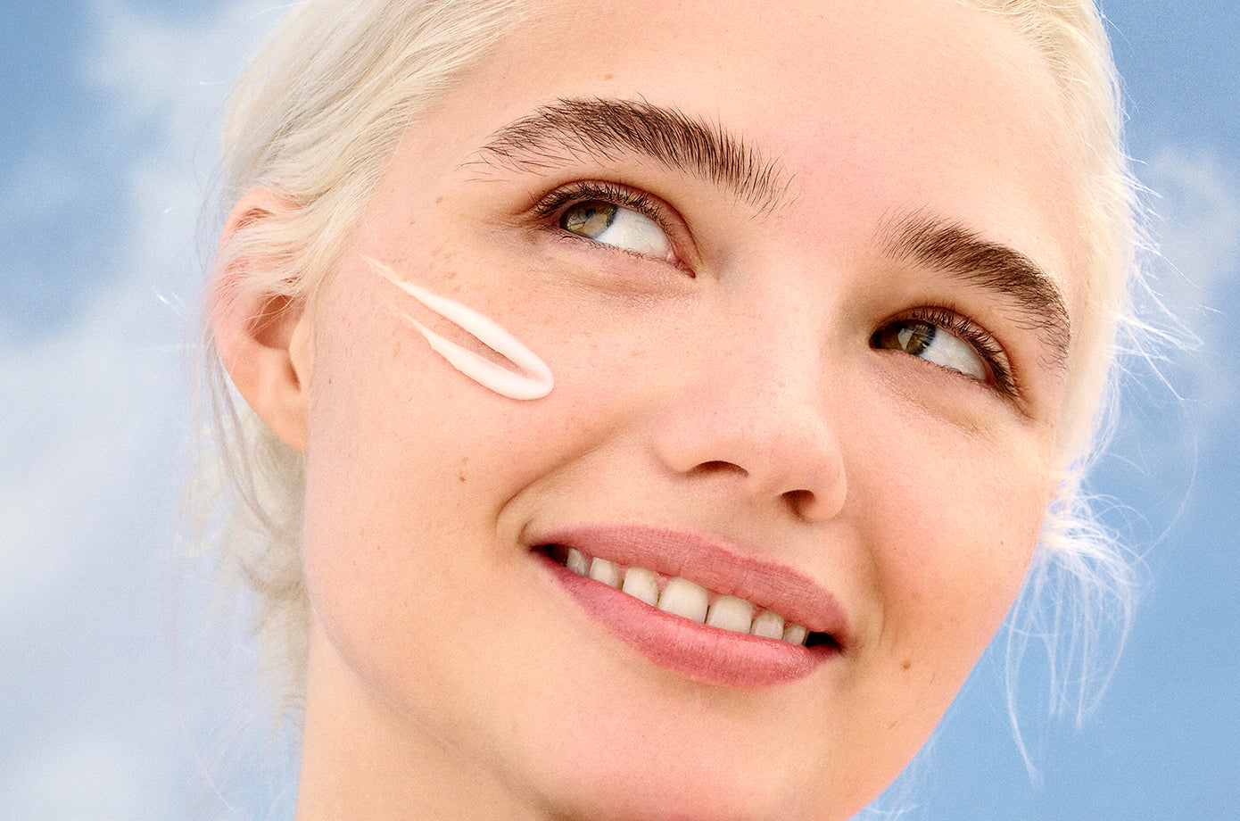 Close-up of a woman applying cream to her face with a blue sky background