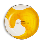 A round ceramic plate with an organic wavy outline, featuring a yellow and white marbled honey-like pattern with the text 'HONEY, YOU'RE GLOWING' printed in black in the center, set against a plain white background