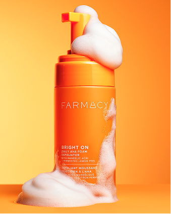 An orange pump bottle of FARMACY Bright On Daily Ana Foam Exfoliator with white foam overflowing from the top and pooling around the base, set against a bright orange background.