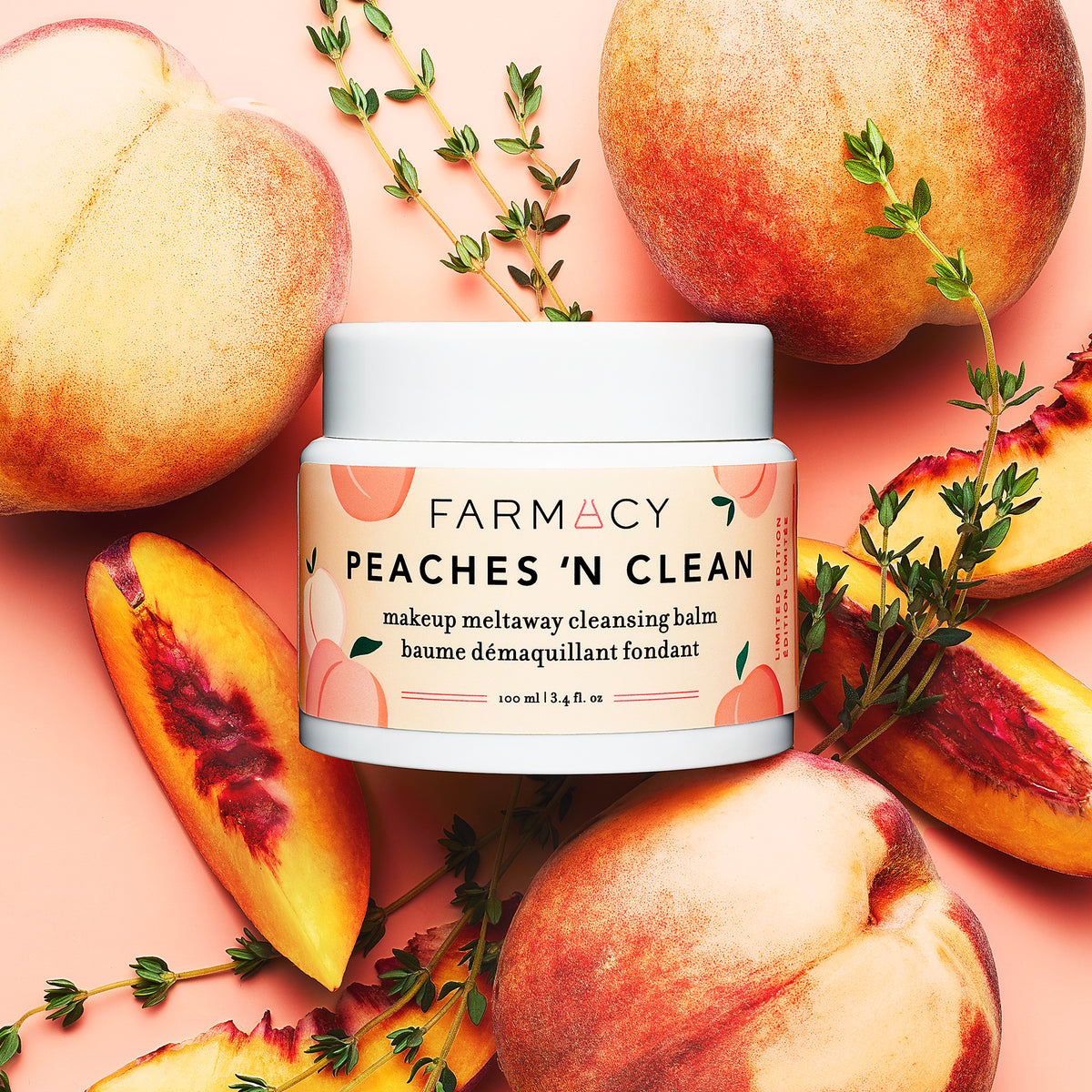 Peaches ’N Clean Makeup Meltaway Cleansing Balm in its closed container surrounded by fresh peaches and herbs on a light pink surface