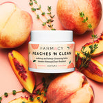 Peaches ’N Clean Makeup Meltaway Cleansing Balm in its closed container surrounded by fresh peaches and herbs on a light pink surface